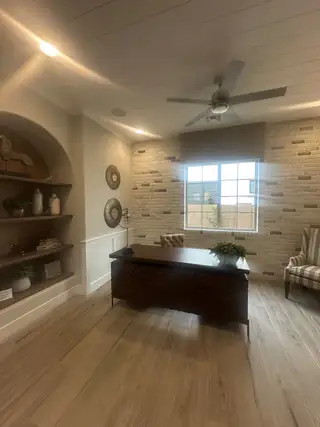 Model Home A modern office with light wood flooring, a textured brick accent wall, cozy chair, and built-in shelves.