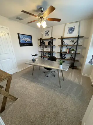Model Home A cozy home office with carpet flooring, modern metal desk, stylish shelves, and ceiling fan, perfect for productivity.