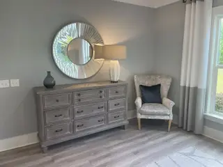 Model Home A cozy corner with a textured gray dresser, chic mirror, soft armchair, and elegant lamp.