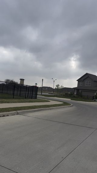 A serene street view in Enchanted Creek by Our Country Homes, Lucas, TX, featuring a modern home under cloudy skies.