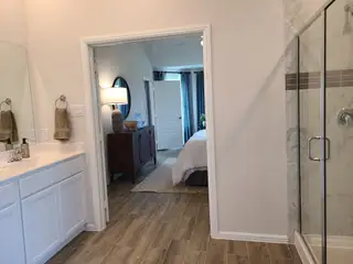 A modern bathroom featuring a glass shower, elegant vanity, and sleek wood flooring leading into a cozy bedroom.
