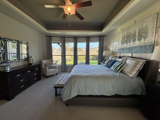 A spacious bedroom with tray ceilings, elegant decor, and large windows offering natural light and a serene view.