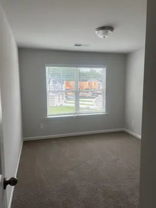 Model Home A bright, carpeted room with a large window and neutral walls, offering a cozy, inviting space.