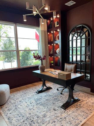 A modern home office with a sleek desk, elegant rug, and built-in shelves, featuring a stylish chandelier and large window.