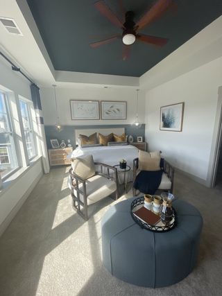 A cozy bedroom with elegant seating, a plush ottoman, and modern artwork, featuring a tray ceiling and ample natural light.