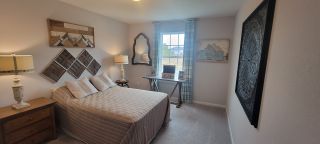 A cozy bedroom with neutral tones, a striped quilt on the bed, decorative wall art, and natural light from a window with patterned curtains.