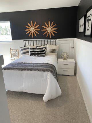 A bedroom with a bold black accent wall, decorative wooden flowers, a metal frame bed with patterned pillows, and white wainscoting.
