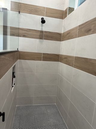 A modern shower featuring wood-patterned and light tiles with a black rain shower head and chic penny tile flooring.