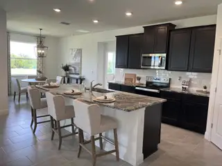 Model Home A modern kitchen with dark cabinets, a granite island, beige seating, and elegant lighting, adjacent to a bright dining area.