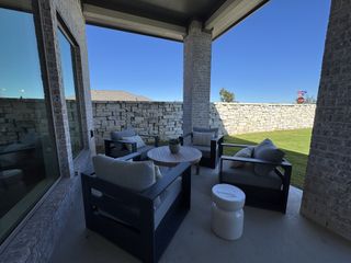 A cozy covered patio with modern seating, neutral tones, and a view of a stone fence and lush lawn. Perfect for relaxation.