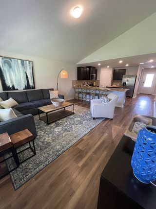 Model Home A spacious living area with a gray sofa, a patterned rug, and hardwood floors flows seamlessly into a modern kitchen with dark cabinets.