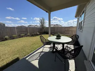A cozy patio with a round table and black wicker chairs overlooks a fenced backyard under a clear blue sky.