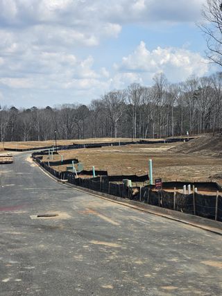 A newly developed street features bare lots with utility connections, bordered by dark fabric fencing and a backdrop of mature trees, in Bailey Fence by Taylor Morrison (Dacula, GA).