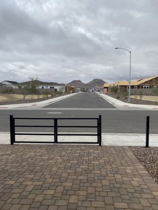 A paved street stretches into the distance, framed by a black metal gate and paver stones, with mountain views under a cloudy sky in Aloravita South by Taylor Morrison (Peoria, AZ).