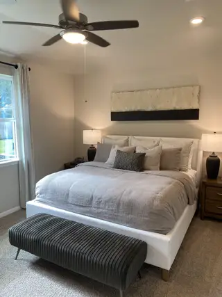 Model Home A cozy bedroom with a plush bed, modern bench, and soft lighting complemented by a ceiling fan and neutral decor.