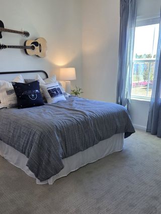 Model Home A cozy bedroom with a plush bed, blue accents, guitar wall decor, and a sunlit window with gray curtains.