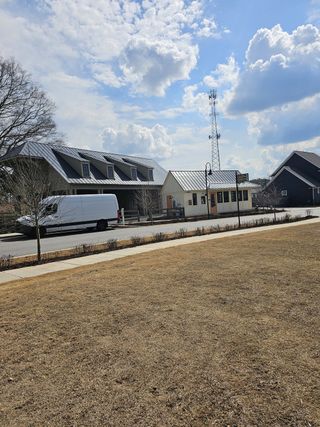 A bright, sunny day showcases a community with modern homes featuring metal roofs, a charming small building with a peach-colored door, and a white van parked along a paved street in Harmony by Kinglett Homes (Auburn, GA).