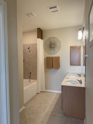 A well-lit bathroom with a double vanity, a soaking tub with a shower curtain, a decorative sunburst mirror, and beige towels, creating a warm and inviting space. 