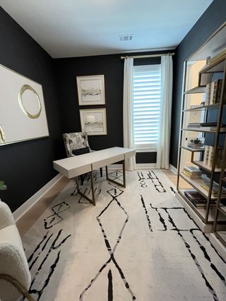 A modern study with a sleek white desk, abstract rug, wall art, and bookshelves, accented by dark walls and soft lighting.