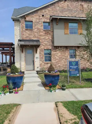 A charming brick home with rustic shutters and flower-lined walkway in The Tribute by Grenadier Homes (The Colony, TX).