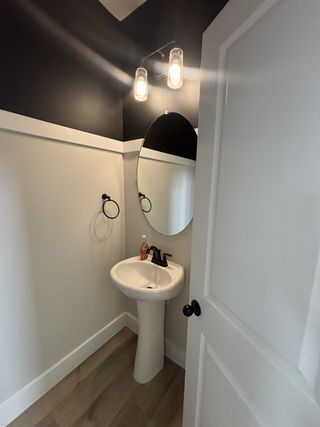 A modern half-bath featuring black and white wall accents, a pedestal sink, and round mirror with stylish lighting.