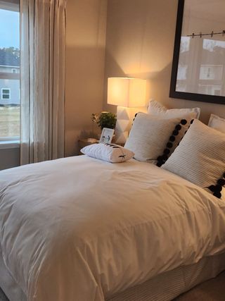 Model Home A cozy bedroom with a plush white comforter, elegant lamp, and a large framed artwork, creating a serene retreat.