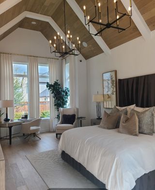 Model Home A luxurious bedroom with vaulted wood ceilings, elegant chandeliers, and cozy seating by the large windows.