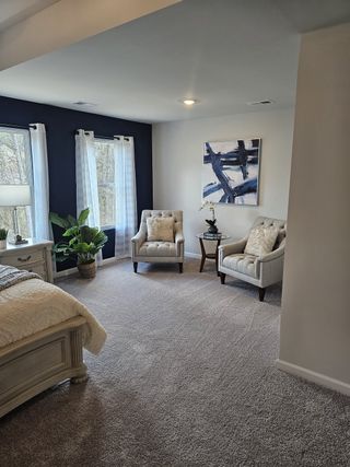 A spacious bedroom features plush carpeting, a dark blue accent wall with large windows, two tufted armchairs, and a decorative abstract artwork, creating a serene and inviting space.