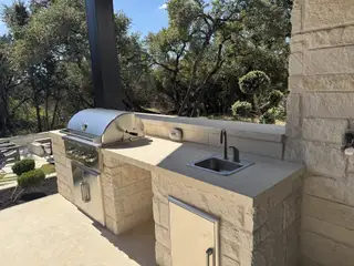 An inviting outdoor kitchen with a sleek grill and sink, set against a serene backdrop of lush trees and natural stone.