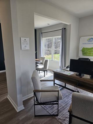 A bright office space features a wooden desk with a computer, a modern armchair, and a glimpse of a dining area with round table and chairs through a doorway.