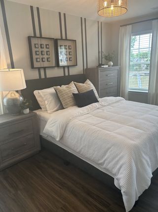 Model Home A cozy bedroom with modern decor, featuring wood floors, a plush bed, chic lighting, and large windows.