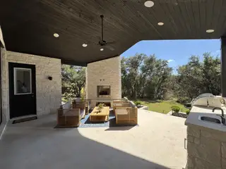 A modern patio with wood seating, a stone fireplace, and outdoor kitchen, offering scenic views of lush greenery.