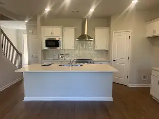 A well-lit kitchen with white shaker cabinets, a center island with a sink, stainless steel appliances, a tiled backsplash, and hardwood floors, adjacent to a staircase.