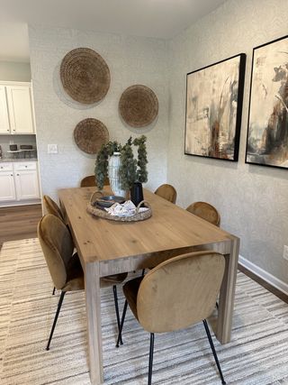 Model Home A modern dining area featuring a wooden table, tan chairs, woven wall decor, and abstract art for a cozy and stylish ambiance.