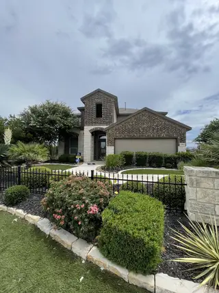 Street view A brick and stone home in Whisper Falls by D.R. Horton (San Antonio, TX) with a two-car garage, black iron fence, and lush landscaping.