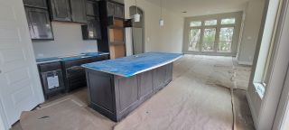 A modern kitchen with dark cabinetry and a large central island, extends into a spacious living area with floor-to-ceiling windows that allow for ample natural light.