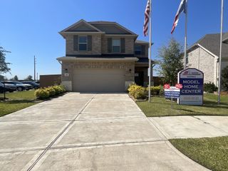 A charming two-story brick home in Sunterra by Adams Homes in Katy with a well-maintained front yard, featuring a model home center sign and promotional banners for zero-down financing.
