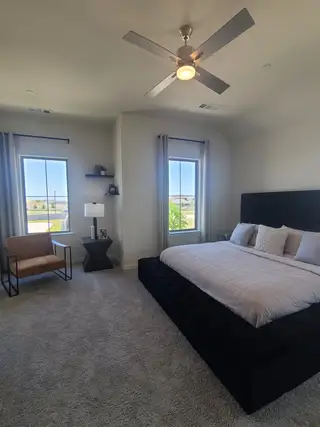 Model Home A cozy bedroom with plush carpet, a modern ceiling fan, and stylish furnishings, complemented by natural light through large windows.