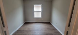 A bright room with soft gray walls, light wood flooring, and a single window offering natural light.