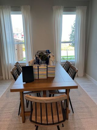 A bright dining area with a wooden table, modern chairs, and sheer white curtains letting in natural light.