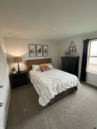 A cozy bedroom with a plush carpet, elegant artwork, and a dark wood dresser, enhanced by soft natural light.