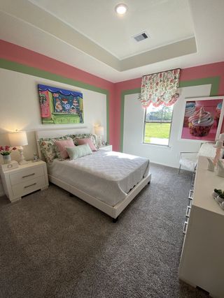 A vibrant bedroom with pink and green accents, decorative art, and floral curtains overlooking a scenic view.