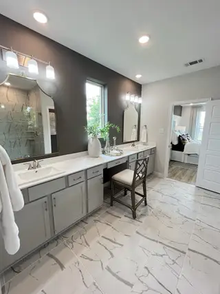 Model Home Elegant bathroom with dual sinks, marble-patterned tiles, chic lighting, and a vanity seating area.
