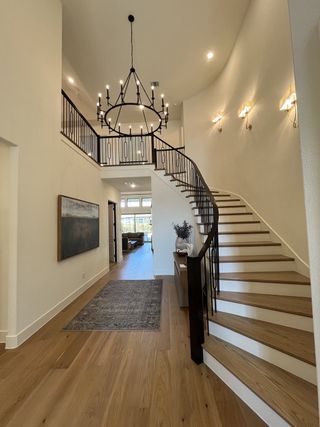 Model Home A grand entrance with a sweeping staircase, elegant chandelier, warm wood floors, and modern wall lighting.