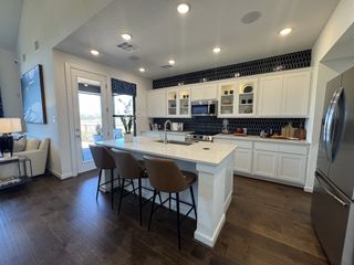 A modern kitchen with sleek white cabinetry, dark backsplash, island seating, and open layout, leading to a cozy living area.