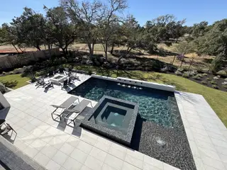 An aerial view of a modern backyard featuring a dark swimming pool with a built-in spa, a fire pit area with seating, and landscaped surroundings.