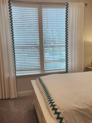 A cozy bedroom featuring white blinds, detailed curtains, and a bed with patterned trim, offering a serene ambiance.