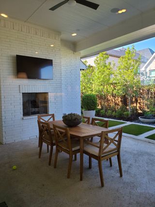 A sleek outdoor patio with a wooden dining set, fireplace, and TV, surrounded by lush greenery.