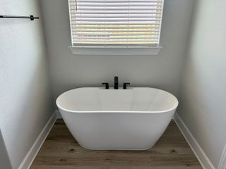 A sleek freestanding bathtub with black fixtures and natural light filtering through blinds, on chic wooden flooring.