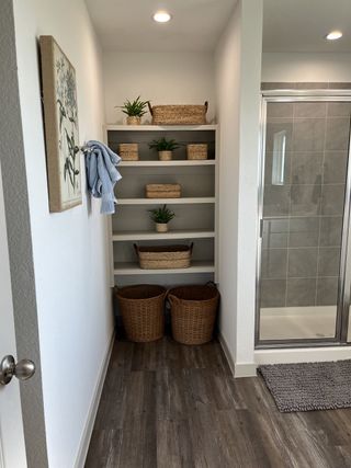 A cozy bathroom nook with woven baskets, plants, and wooden flooring, adjacent to a sleek glass shower.